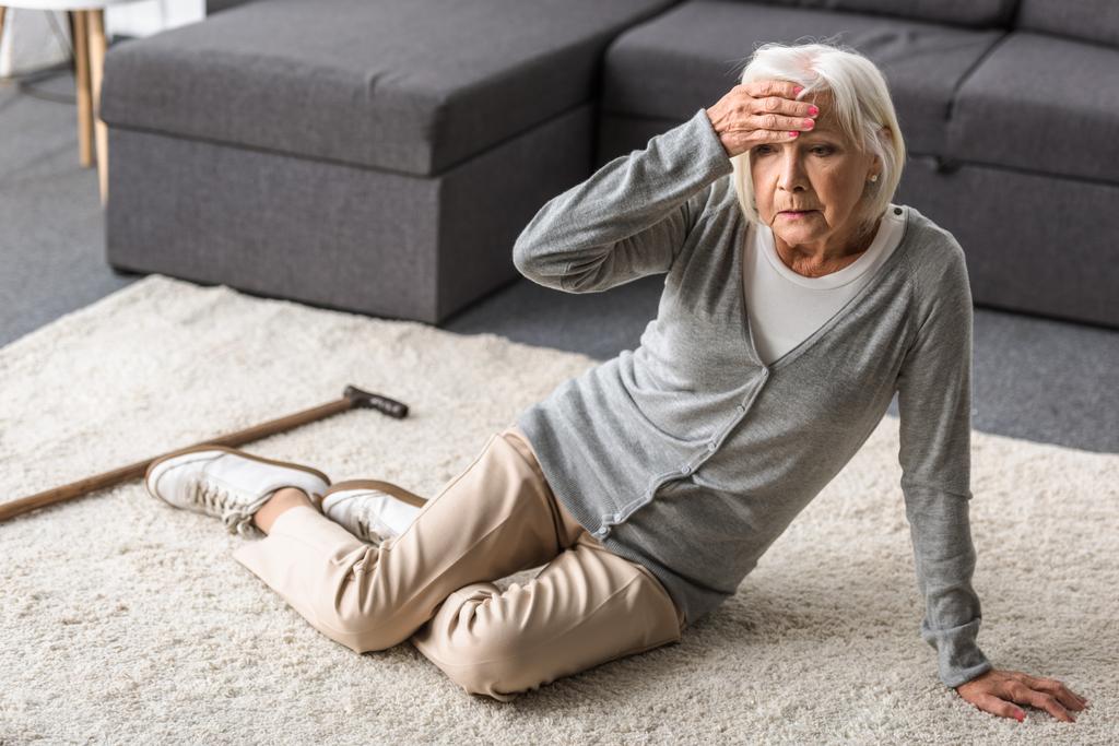 Seniorin mit Migräne sitzt auf Teppich und berührt Stirn mit der Hand - Foto, Bild