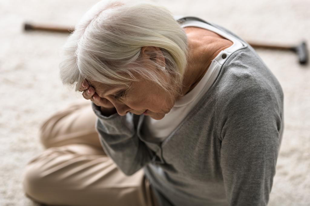 Seniorin mit Migräne sitzt auf Teppich und berührt Stirn mit der Hand - Foto, Bild