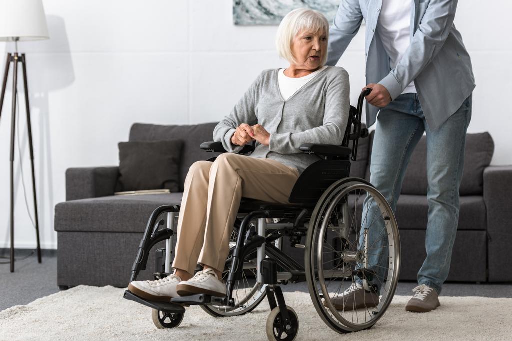 Ausgeschnittener Blick auf Mann, der Seniorin im Rollstuhl trägt - Foto, Bild