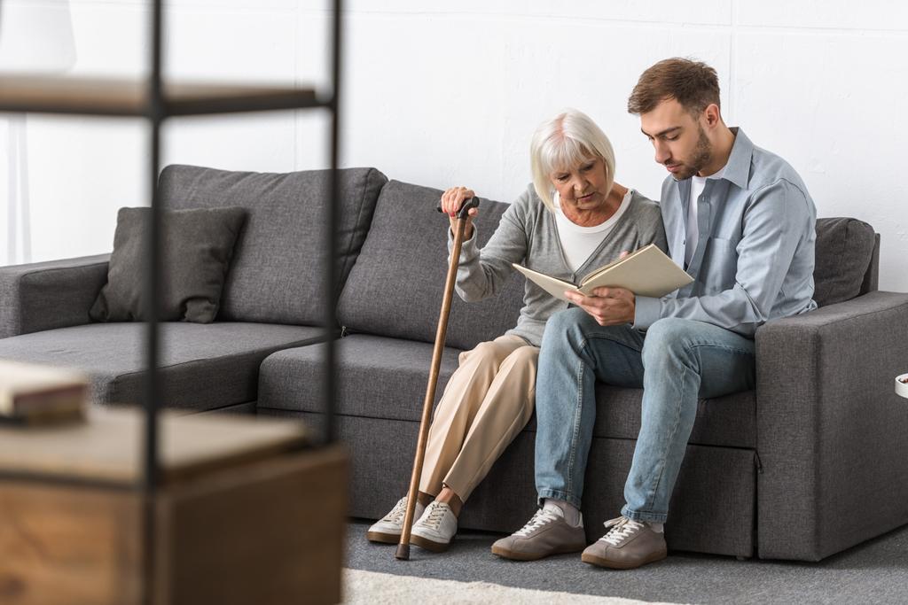 Mann sitzt mit älterer Mutter auf Sofa und liest Buch - Foto, Bild