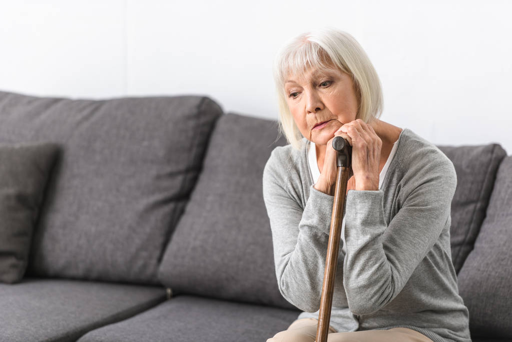 Nachdenkliche Seniorin mit Stock auf Sofa sitzend und nach unten blickend - Foto, Bild
