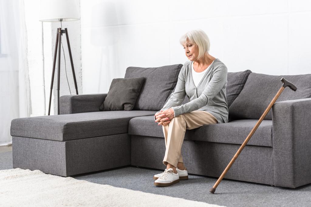 pensive vanhempi nainen puinen keppi istuu sohvalla olohuoneessa
 - Valokuva, kuva