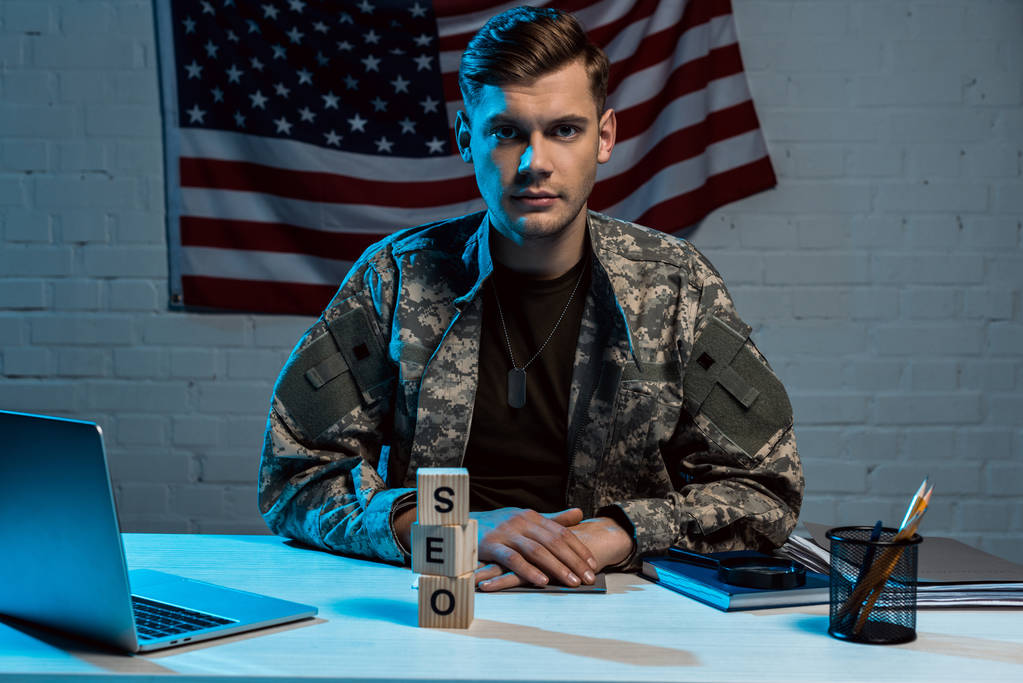 красивий військовий чоловік сидить зі згорнутими руками біля ноутбука та кубиків з сео-нотацією
  - Фото, зображення