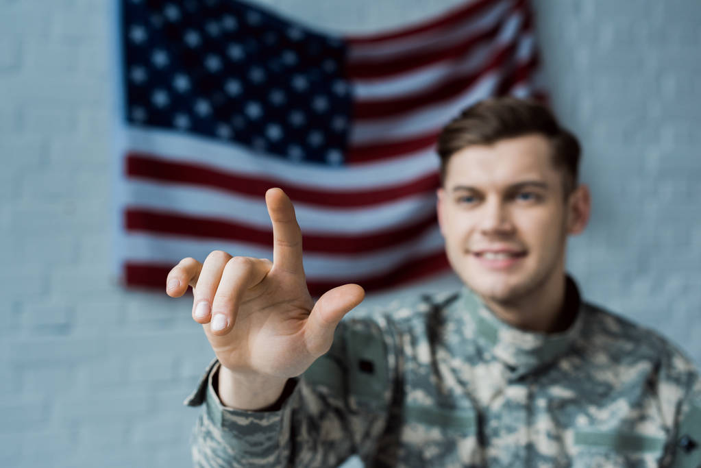 selektywne skupienie szczęśliwego człowieka w mundurze wojskowym wskazując palcem w pobliżu amerykańskiej flagi - Zdjęcie, obraz