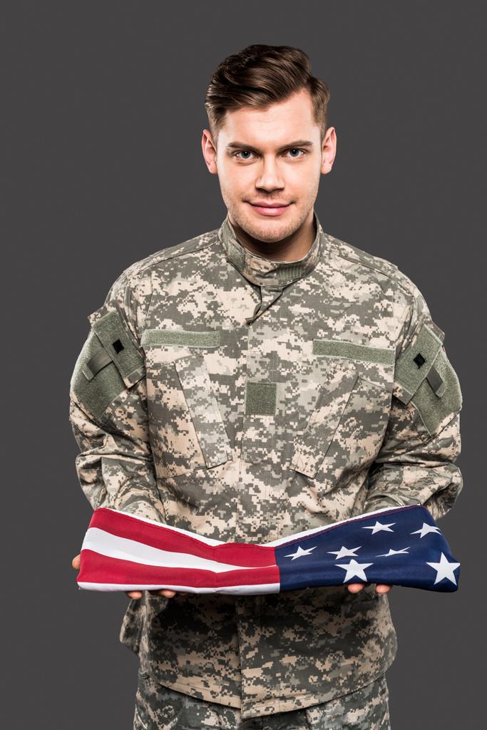 ευτυχισμένος άνθρωπος με στρατιωτική στολή κρατώντας αμερικάνικη σημαία απομονωμένη σε γκρι  - Φωτογραφία, εικόνα