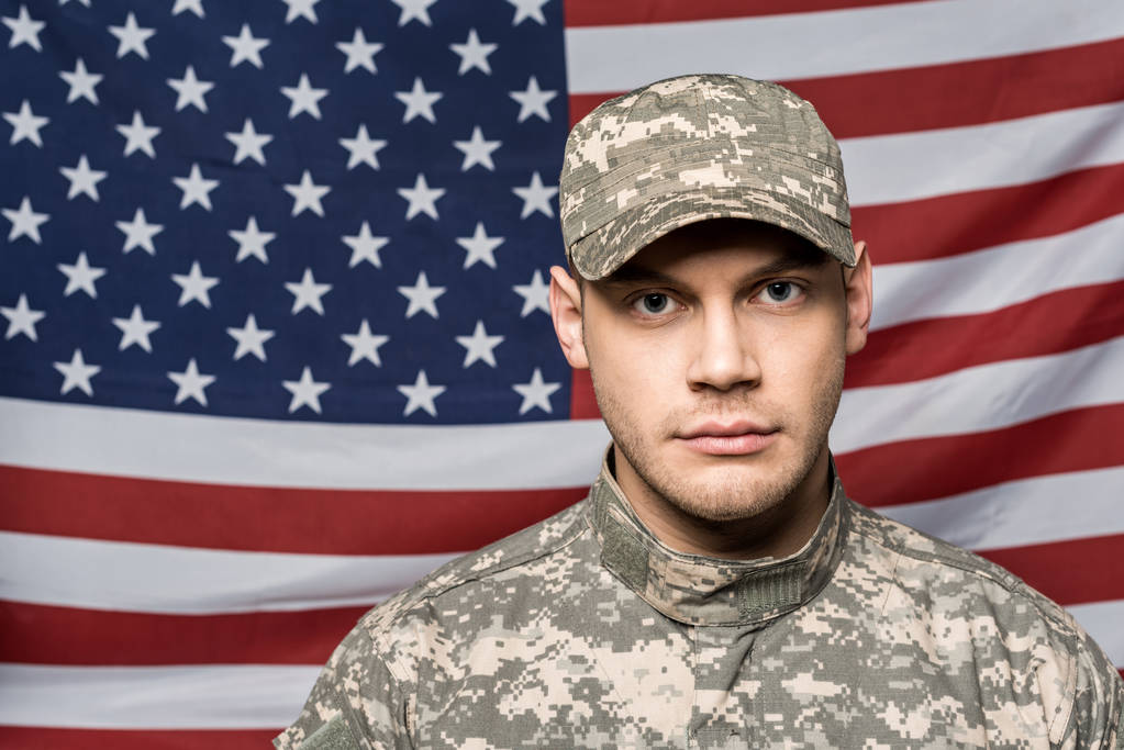 schöner Soldat in Militäruniform und Mütze, der in die Kamera neben der Flagge Amerikas blickt - Foto, Bild