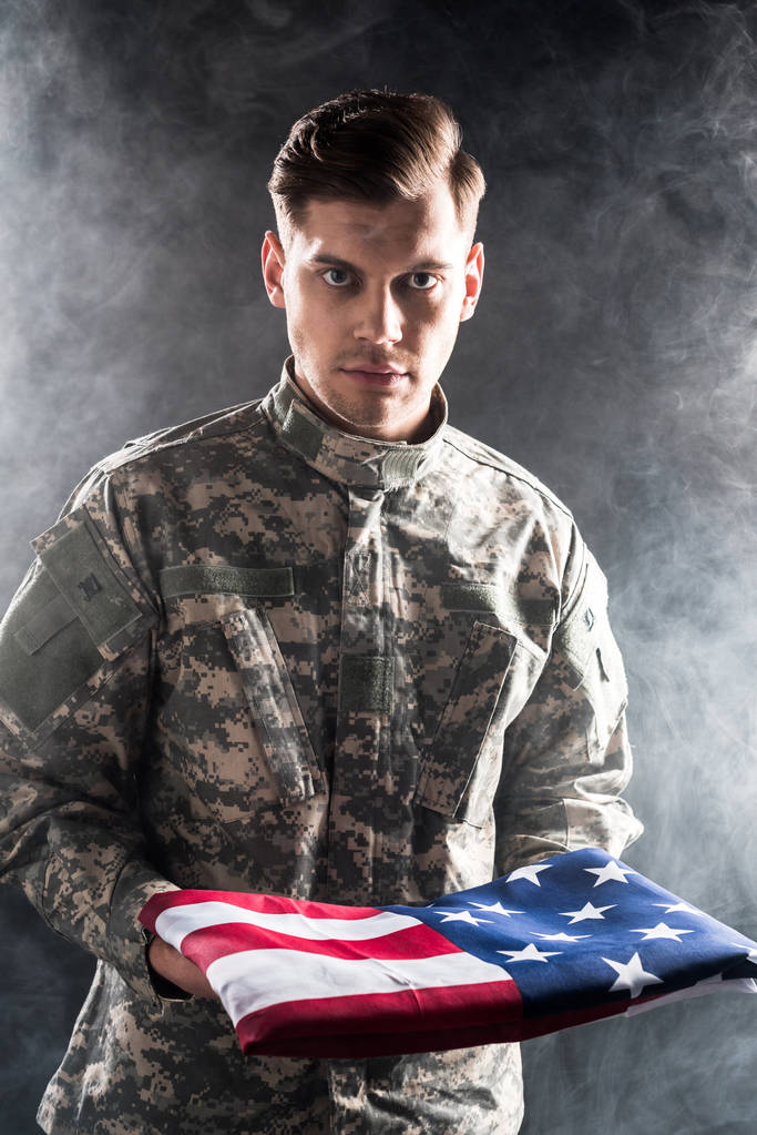 knappe man in militair uniform houden Amerikaanse vlag op zwart met rook  - Foto, afbeelding