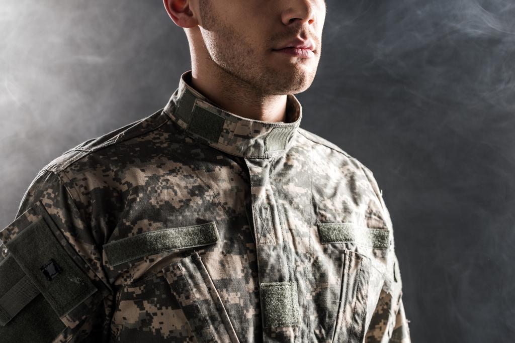 bijgesneden weergave van serieuze soldaat in militair uniform op zwart met rook  - Foto, afbeelding