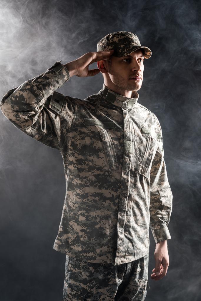 soldier in military uniform and cap giving salute on black with smoke - Photo, Image
