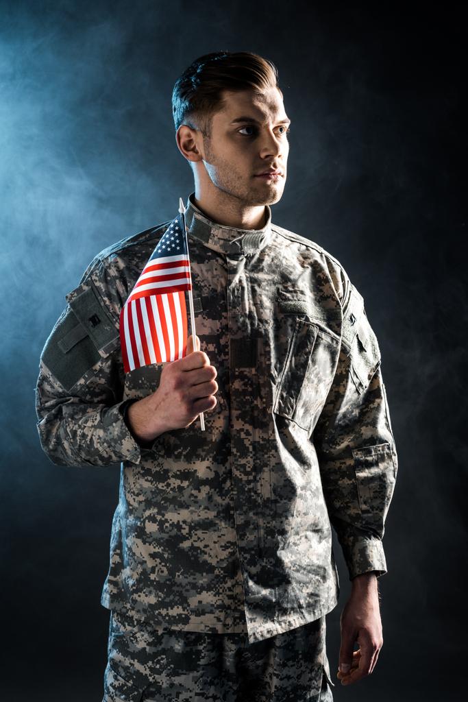 στρατιώτης με αυτοπεποίθηση σε στρατιωτική στολή, κρατώντας την αμερικάνικη σημαία σε μαύρο με καπνό  - Φωτογραφία, εικόνα