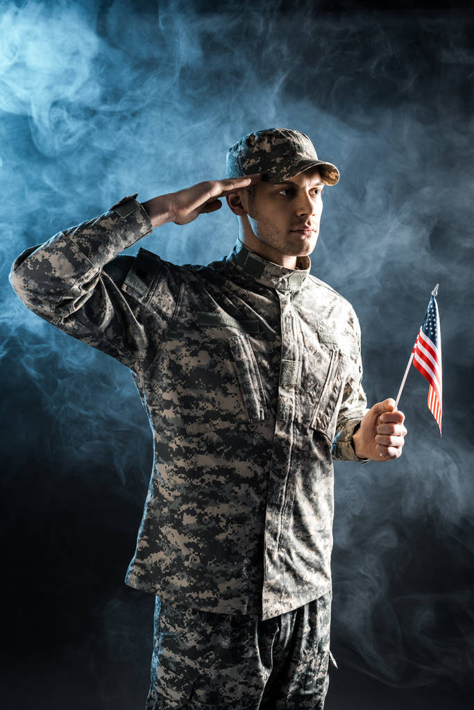 Selbstbewusster Soldat in Militäruniform mit amerikanischer Flagge und Salutschüssen auf schwarz mit Rauch  - Foto, Bild