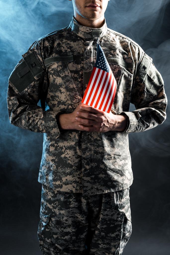 煙で黒い上にアメリカの旗を保持する迷彩制服の男のトリミングされたビュー  - 写真・画像