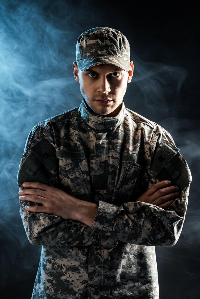 zelfverzekerde soldaat in militair kapje en uniform staand met gekruiste armen op zwart met rook - Foto, afbeelding
