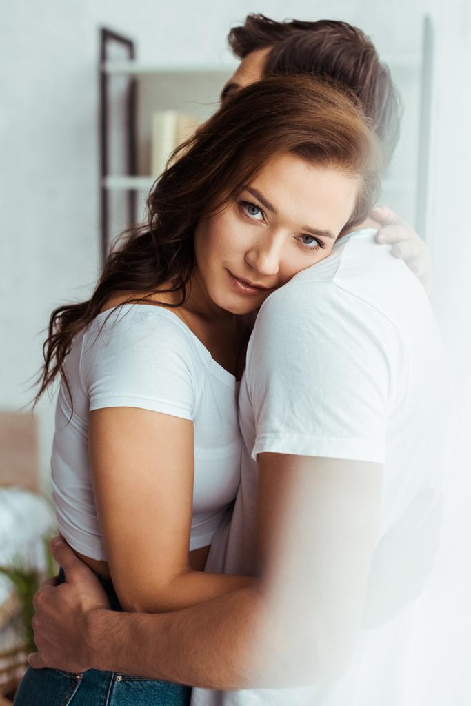 Freund umarmt attraktive Freundin mit Blick auf Kamera zu Hause   - Foto, Bild