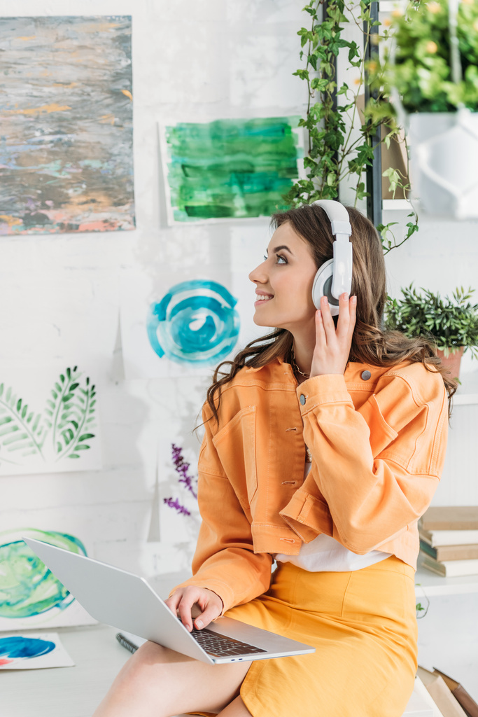 Gelukkige vrouw kijkt naar schilderijen op de muur tijdens het luisteren van muziek in een koptelefoon en het gebruik van laptop - Foto, afbeelding
