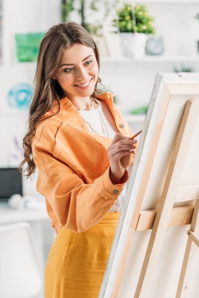 cheerful young artist in trendy clothing painting on easel at home - Photo, Image