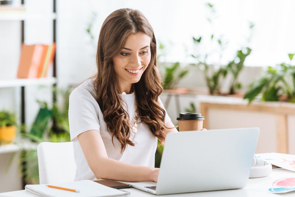 alegre joven mujer usando el ordenador portátil y sosteniendo la taza de papel mientras está sentado en el escritorio en casa
 - Foto, Imagen