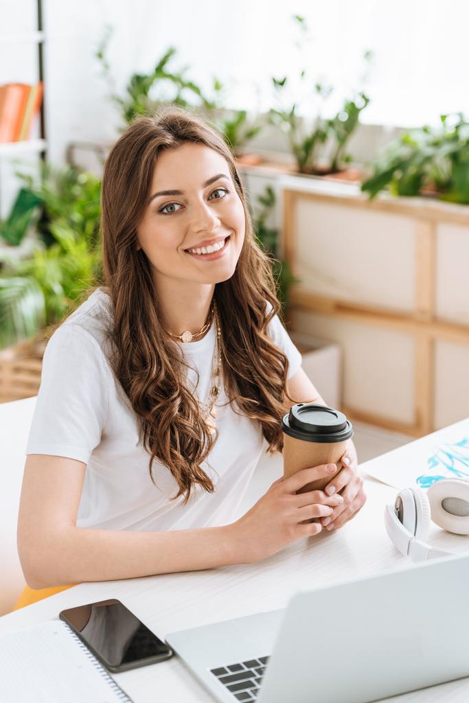 χαρούμενη νεαρή γυναίκα κοιτάζοντας την κάμερα και κρατώντας κύπελλο χαρτί ενώ κάθεται στο γραφείο με φορητό υπολογιστή - Φωτογραφία, εικόνα