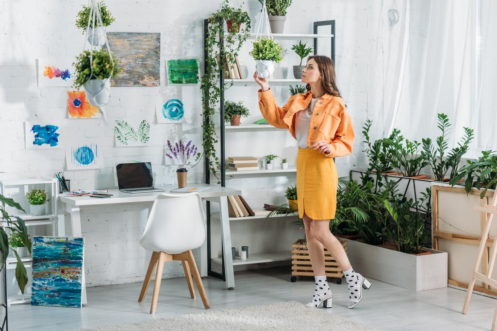 trendikäs nuori nainen koskettaa kukkaruukku seisoessaan tilava huone maalauksia seinään, telineeseen ja ruukkukasveja
 - Valokuva, kuva