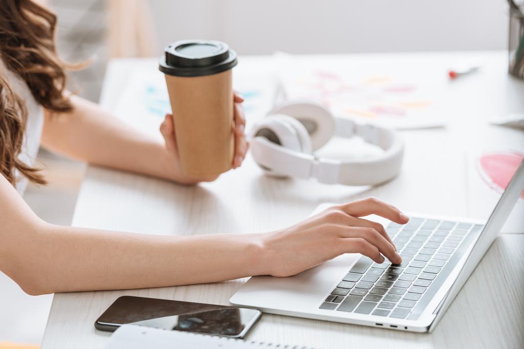 cropped view of young woman holding paper cup while using laptop near headphones - Photo, Image
