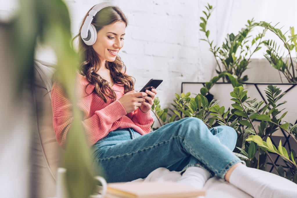 selectieve focus van glimlachend meisje met smartphone terwijl je zit omringd door groene planten en luister muziek in een koptelefoon - Foto, afbeelding