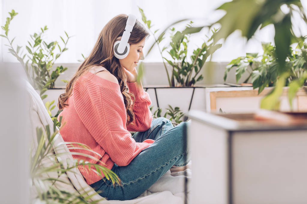 mujer joven pensativa escuchando música en auriculares mientras está sentada rodeada de plantas verdes en casa
 - Foto, Imagen