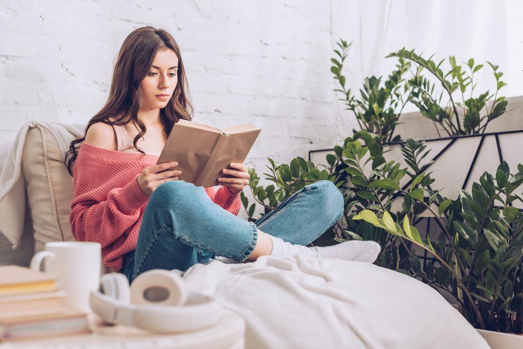 messa a fuoco selettiva di focalizzata giovane donna lettura libro mentre seduto con le gambe incrociate vicino piante verdi a casa
 - Foto, immagini