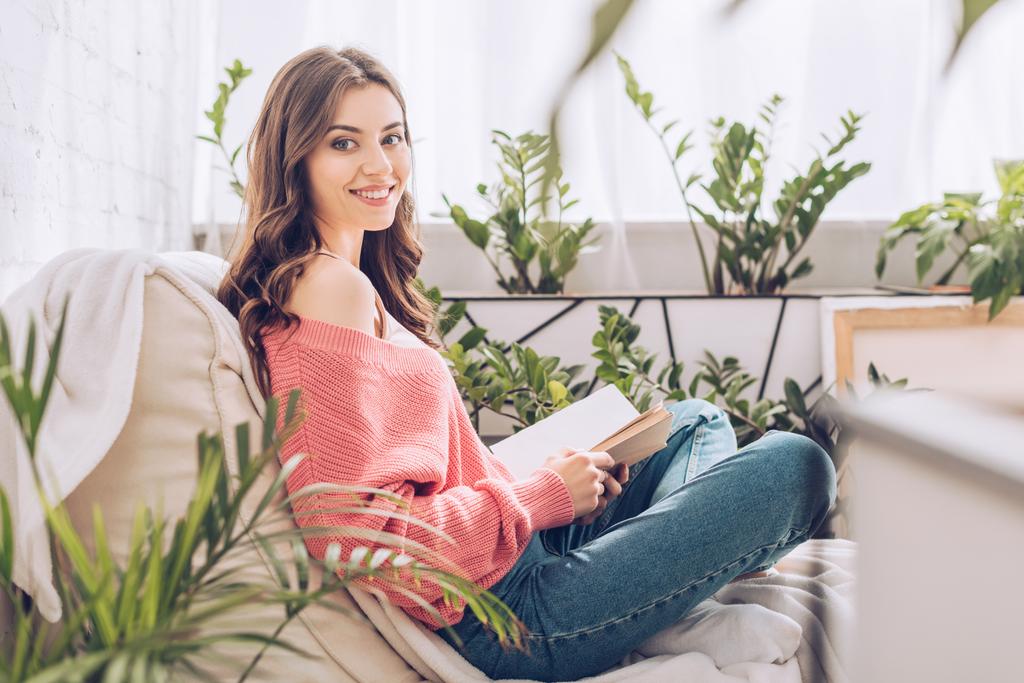 szelektív középpontjában a vidám lány, kezében könyvet, és mosolyogva a kamera mellett ül a szobában zöld növényekkel  - Fotó, kép