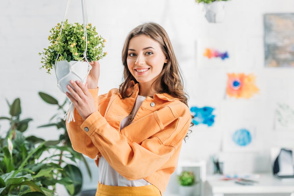 vrolijke vrouw aanraken opknoping bloempot met groene plant en glimlachend op de camera - Foto, afbeelding