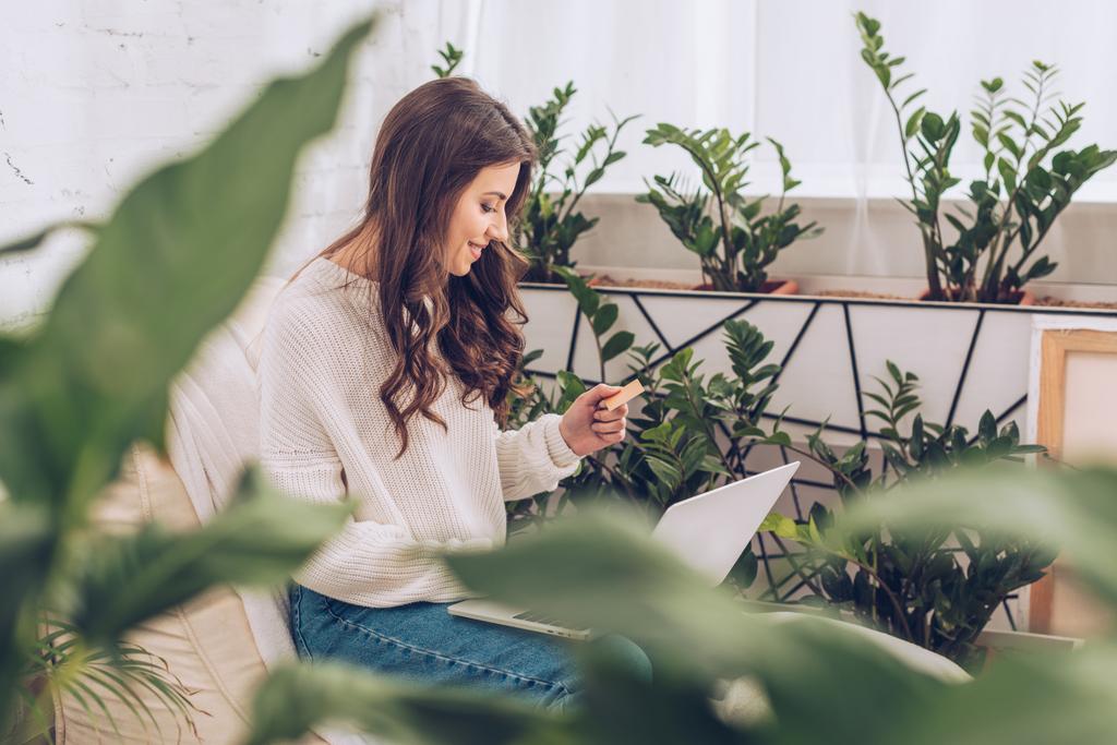 自宅で緑の植物に囲まれて座っている間、クレジットカードを持ち、ノートパソコンを使用して笑顔の女性の選択的な焦点 - 写真・画像