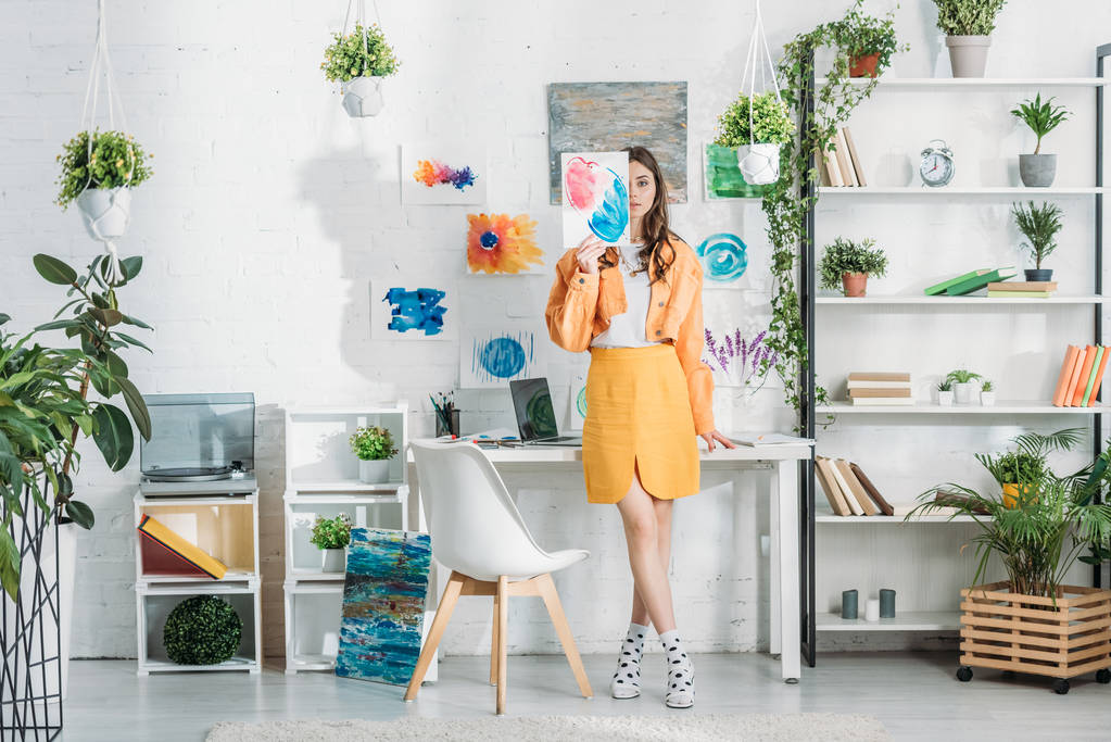 jovem mulher na moda cobrindo rosto com pintura enquanto está em pé em quarto espaçoso decorado com plantas verdes e desenhos na parede branca
 - Foto, Imagem