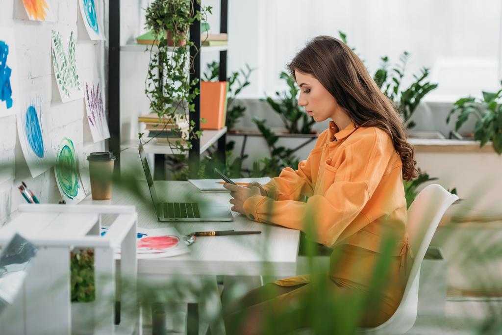 Selektiver Fokus der nachdenklichen jungen Frau, die am Schreibtisch neben dem Laptop sitzt und das Smartphone nutzt - Foto, Bild