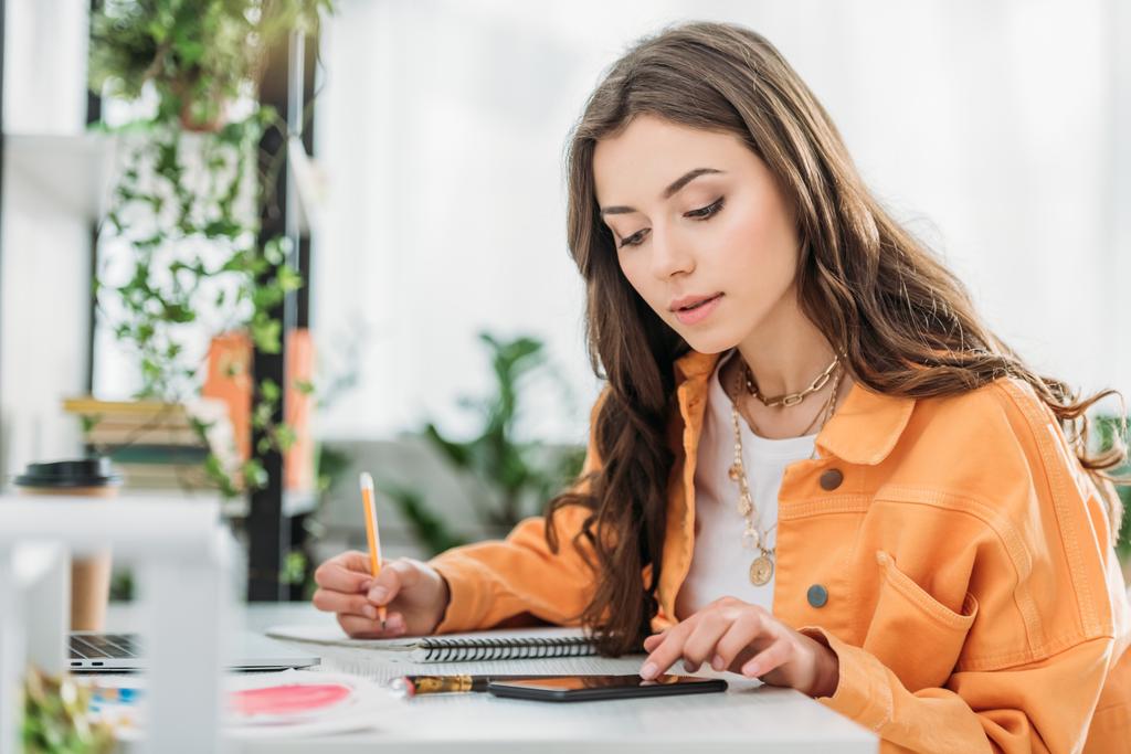 προσεκτική νεαρή γυναίκα που κάθεται στο γραφείο, χρησιμοποιώντας smartphone και γράφοντας στο σημειωματάριο  - Φωτογραφία, εικόνα