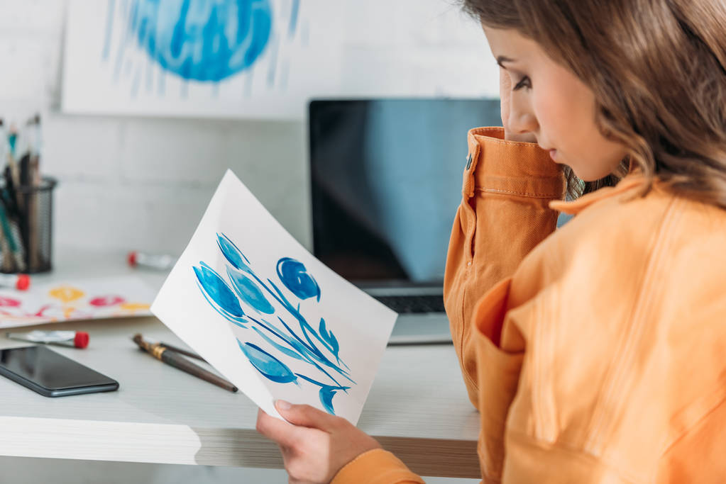 σκεπτική νεαρή γυναίκα κάθεται στο γραφείο και κοιτάζοντας τη ζωγραφική με μπλε λουλούδια - Φωτογραφία, εικόνα