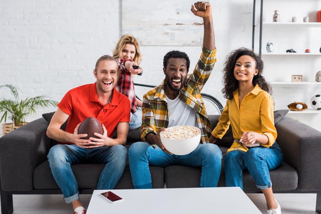 Šťastní multikulturní přátelé sedí na gauči a sledují šampionát u stolu s popcornem v misce - Fotografie, Obrázek