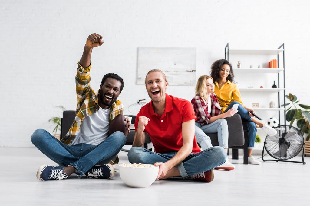Счастливые и позитивные мультикультурные молодые люди сидят на полу и смотрят спортивную игру с миской попкорна
 - Фото, изображение