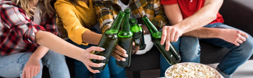 Περικομμένη θέα τεσσάρων πολυεθνικών φίλων με μπουκάλια μπύρας που ξοδεύουν χρόνο μαζί στο καθιστικό - Φωτογραφία, εικόνα
