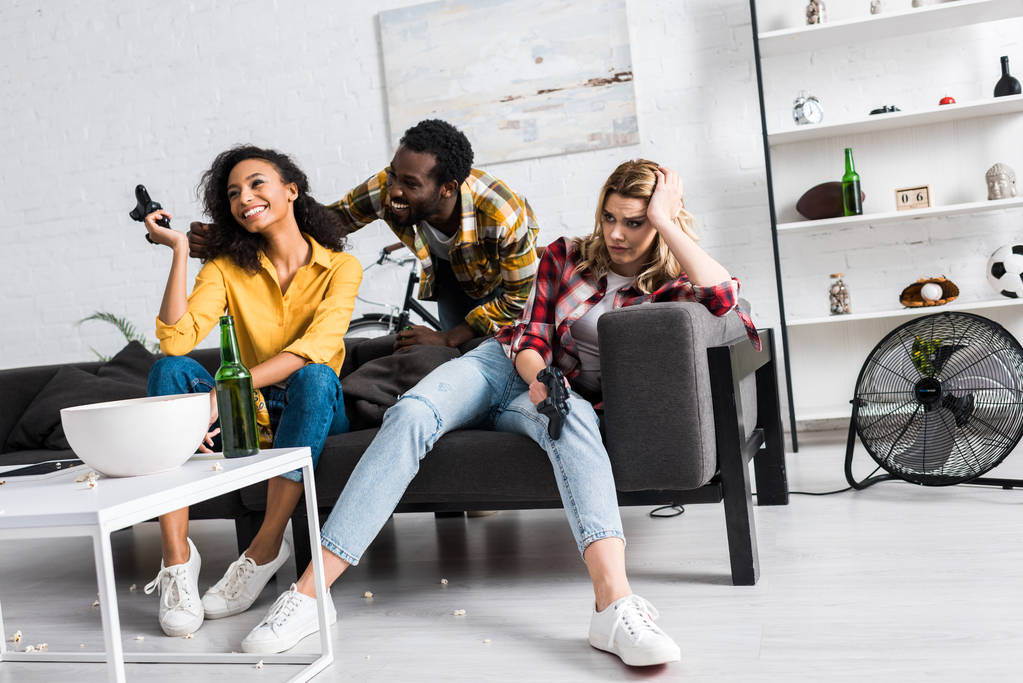 vista basso angolo di ragazza sconvolto seduto vicino amici afro-americani felici in soggiorno
  - Foto, immagini
