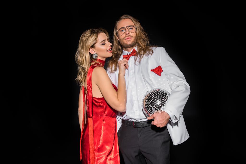 aantrekkelijke vrouw in rode jurk aanraken Bow tie van de mens in glazen met disco bal geïsoleerd op zwart  - Foto, afbeelding