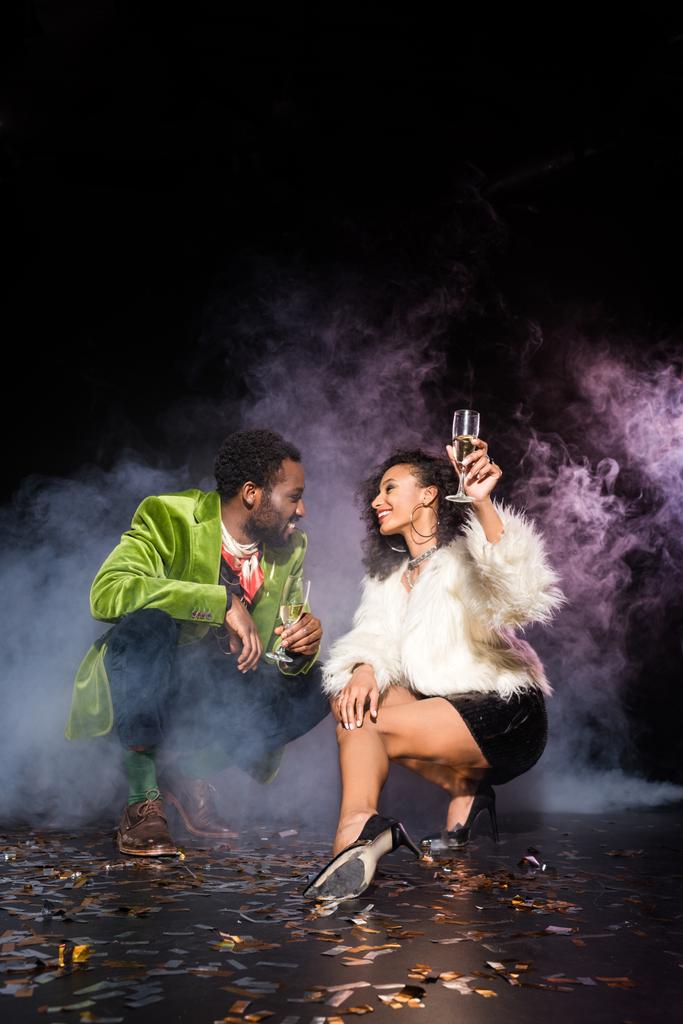 afroamericano uomo e donna in possesso di bicchieri di champagne mentre si guardava vicino coriandoli sul nero con il fumo
  - Foto, immagini