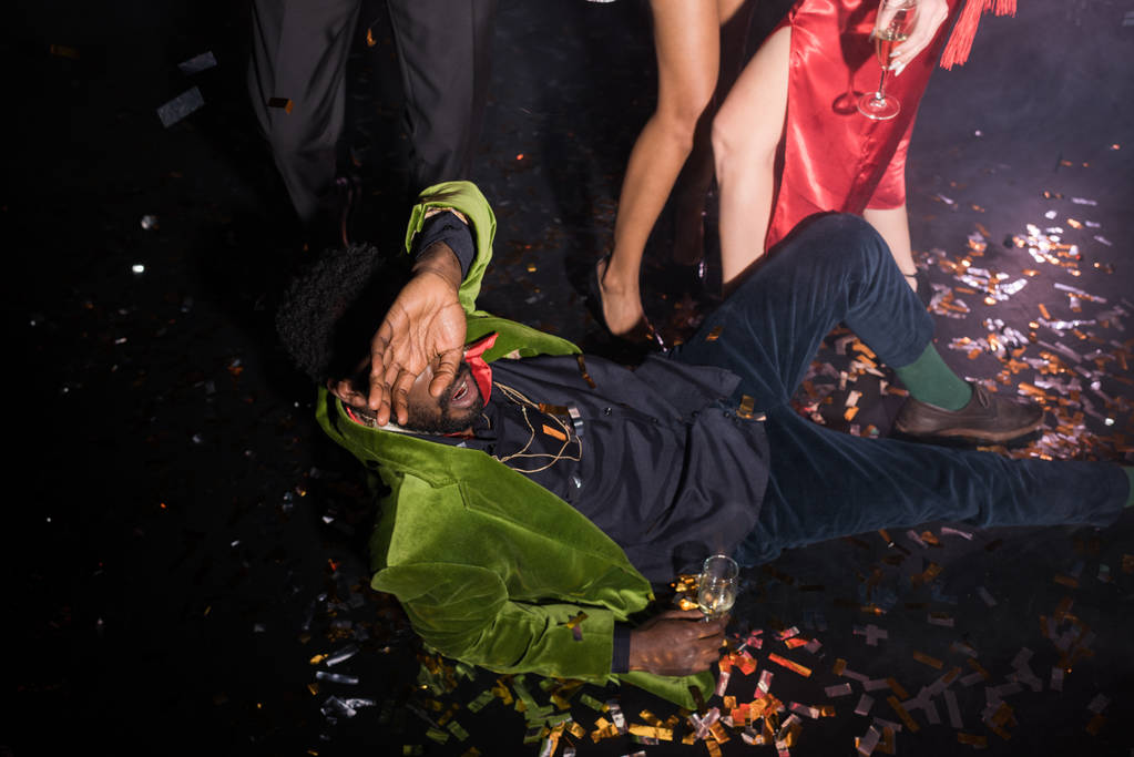 pijany African American Man pokrycie twarzy podczas leżącego na podłodze z konfetti w pobliżu przyjaciół na czarnym  - Zdjęcie, obraz