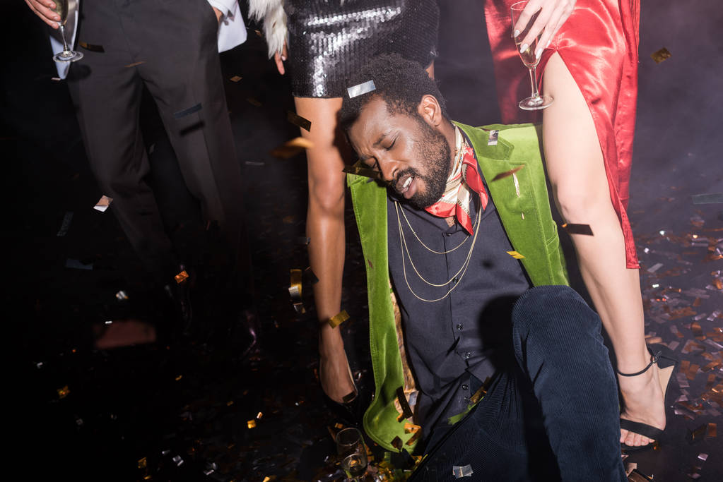 borracho afroamericano hombre con los ojos cerrados acostado en el suelo con confeti brillante cerca de amigos en negro
  - Foto, imagen