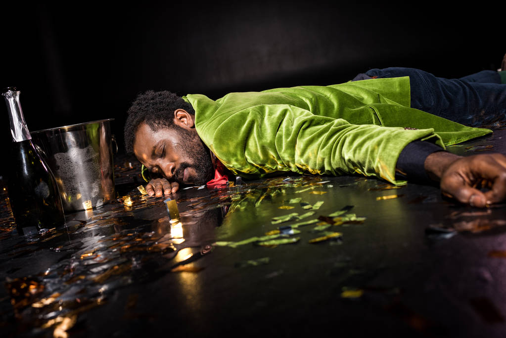氷のバケツの近くに紙吹雪と黒のシャンパンのボトルで床に横たわっている酔ったアフリカ系アメリカ人男性の選択的な焦点  - 写真・画像