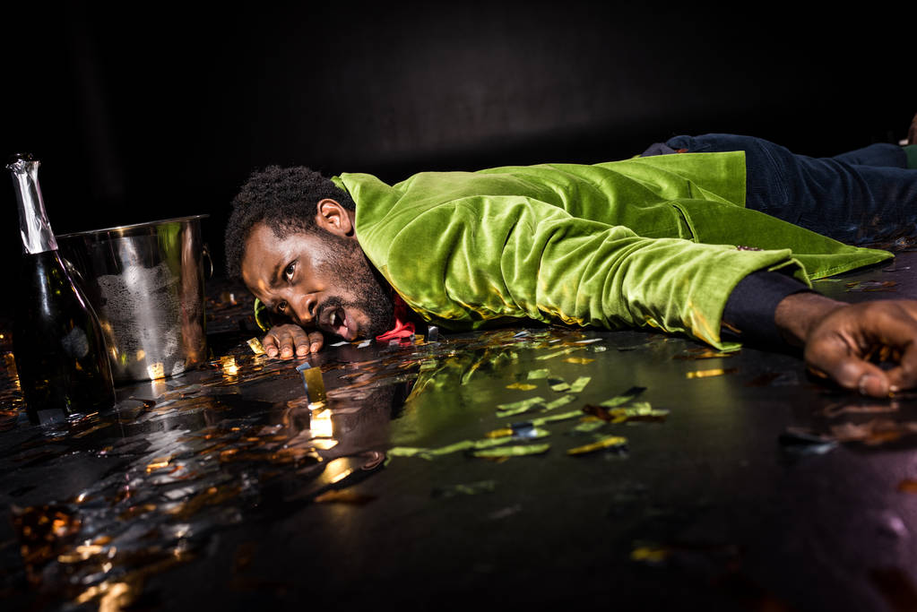 szelektív összpontosít részeg afro-amerikai ember feküdt a padlón fényes konfetti közelében jég vödör és üveg pezsgő fekete  - Fotó, kép