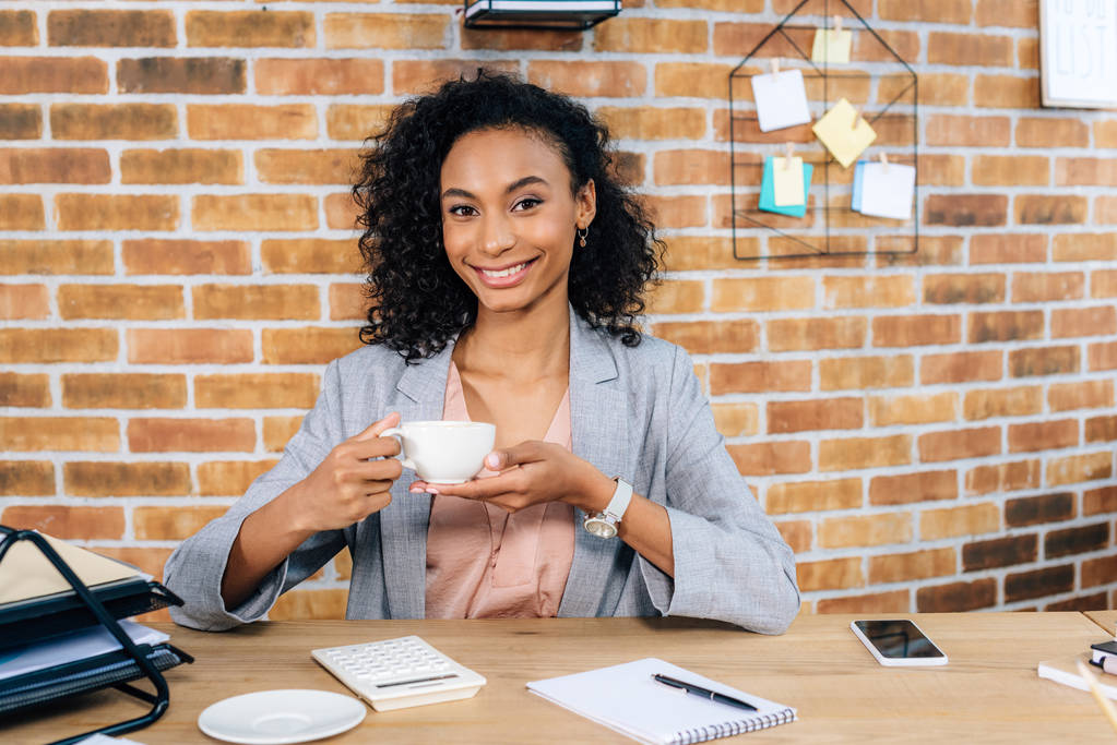 オフィスでコーヒーカップを持つ机で笑顔のアフリカ系アメリカ人カジュアルビジネスウーマン - 写真・画像
