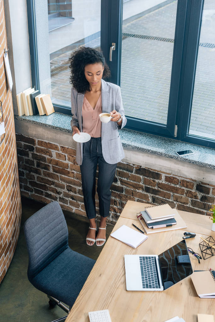 красива афроамериканка Випадкова бізнес-леді з чашкою кави біля офісного столу з ноутбуком
 - Фото, зображення