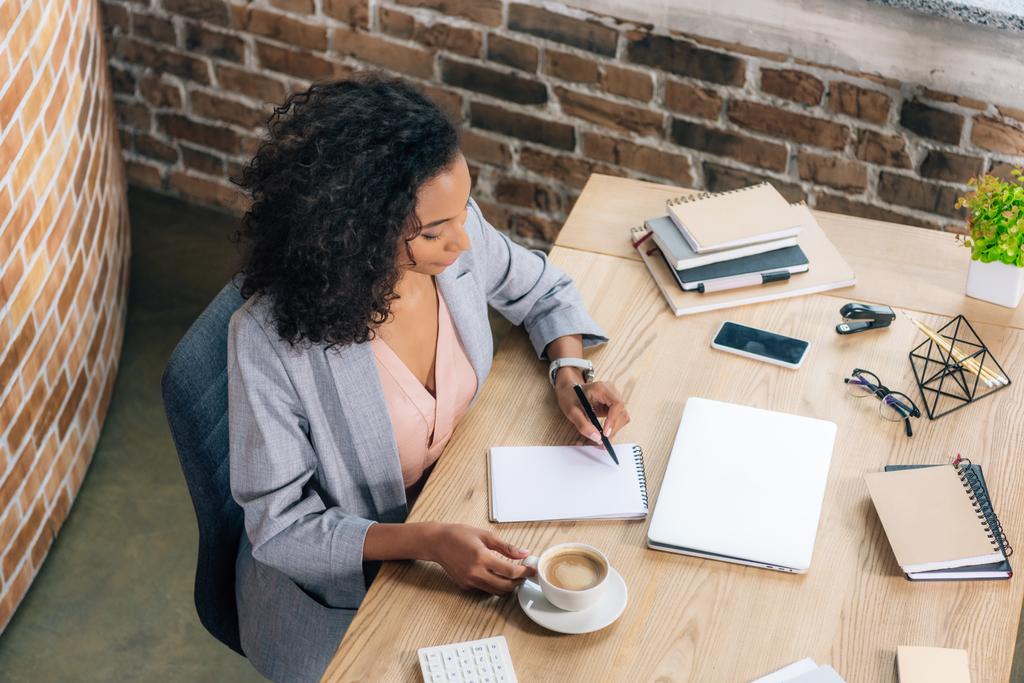 Casual mulher de negócios afro-americana sentada na mesa e escrevendo em notebook no escritório loft
 - Foto, Imagem