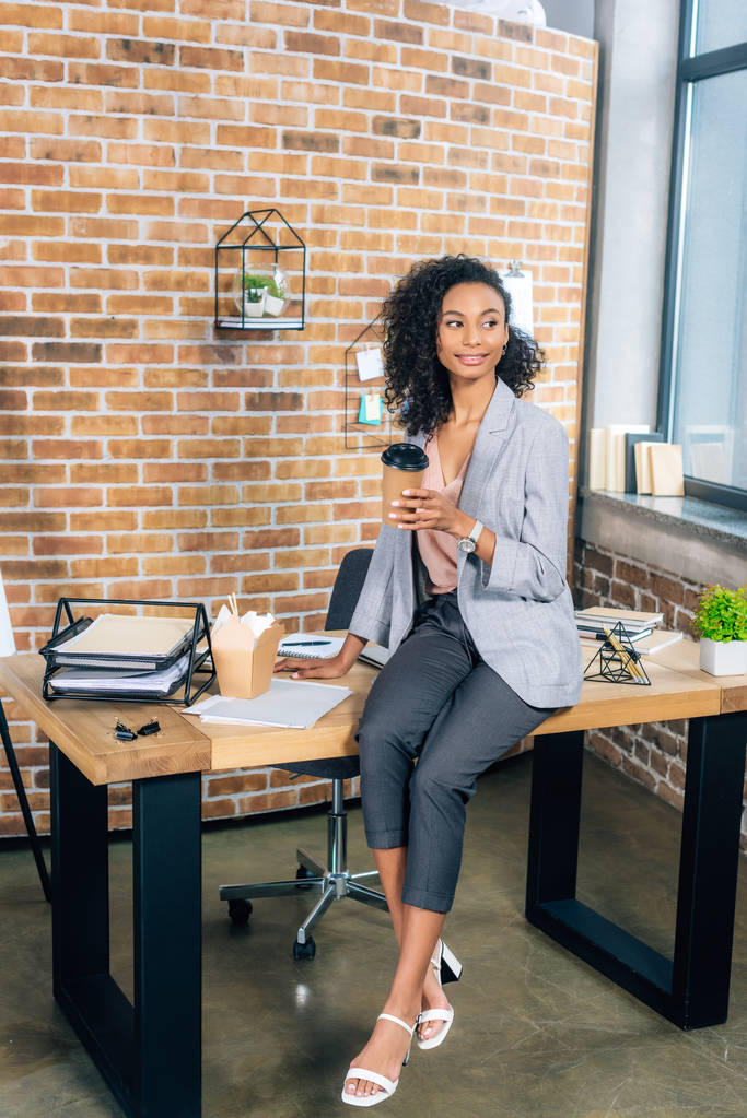 Afrikanisch-amerikanische Gelegenheitsunternehmerin sitzt am Schreibtisch mit Coffee to go und Imbisskasse im Büro - Foto, Bild