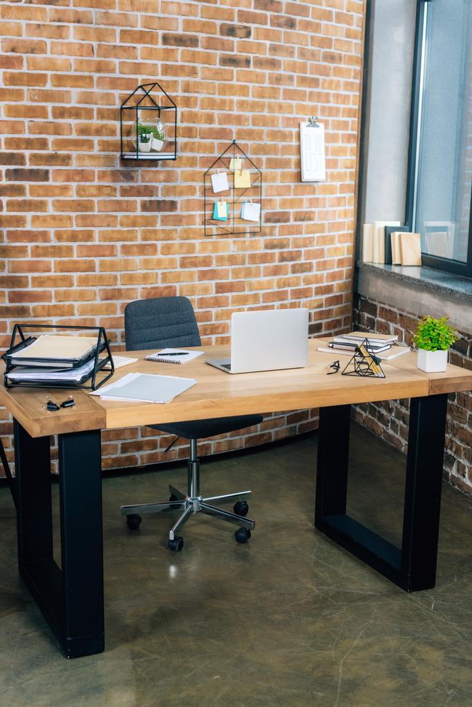 письменный стол с ноутбуком и документы в современном лофт-офисе с кирпичной стеной
 - Фото, изображение