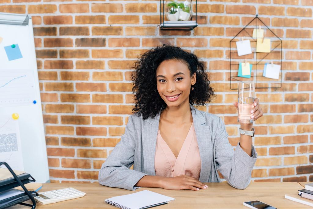 usmívající se africká americká příležitostná Obchodná žena, která drží sklenici vody u kancelářského stolu - Fotografie, Obrázek